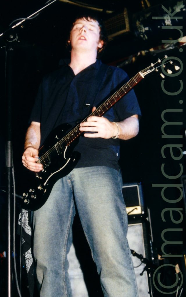 Low view of a man with dark hair, wearing a black shirt and a faded blue pair of jeans, playing a black electric guitar. He is leaning back slightly, with his eyes closed and his mouth slightly open. Black and grey amplifiers line the black walls in the background, with the ceiling also being black.