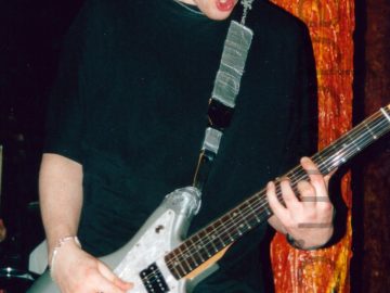A man with short dark hair and wearing a black t-shirt and a blue pair of jeans, and playing a metallic-grey and white electric guitar. A black guitar strap, covered in grey tape, is attached to the top and bottom of the guitar, and slung over his left shoulder. He is looking down at his instrument, his mouth slightly agape. A brown, orange, and yellow pillar, looking almost organic, is behind his left arm, stretching from top to bottom of the frame. Dark walls and ceiling fill the rest of the frame, with green stagelights and yellow room lights visible here and there.