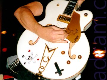 Closeup of a white electric guitar. The hands and arms of the person holding it are visible, but most of his body is hidden behind it as he leaps into the air, his feet in black and red shoes with white socks being the only other part of him that sticks out. Black walls with orange stage lights fill the rest of the frame.