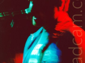 A man standing on a stage, singing into a microphone on a stand in front of him. The photo is taken with a long exposure, with stage lights flooding various parts of the frame with different vivid colours, including green on his t-shirt and face and the microphone, blue on his jacket, and red on his back and hair. Black walls fill the background.