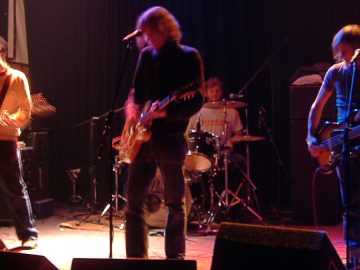 Four members of a band performing on a dark, atmospherically lit stage. On the left is a man wearing a black knitted hat, a white jumper, and dark trousers, playing a black and white electric guitar. In the middle is a man with long, wavy blond hair, wearing a black jumper and dark trousers, playing a brown electric guitar. Behind him is a wan with short dark hair, wearing a white t-shirt and sitting behind a black drum kit. On the right is a man with long, straggly blond hair, wearing a dark t-shirt and blue jeans, and playing a black and brown electric bass guitar. Black curtains fill the background, behind black and grey amplifiers. A long exposure has been used, with movement of heads, arms, and instruments being visible.