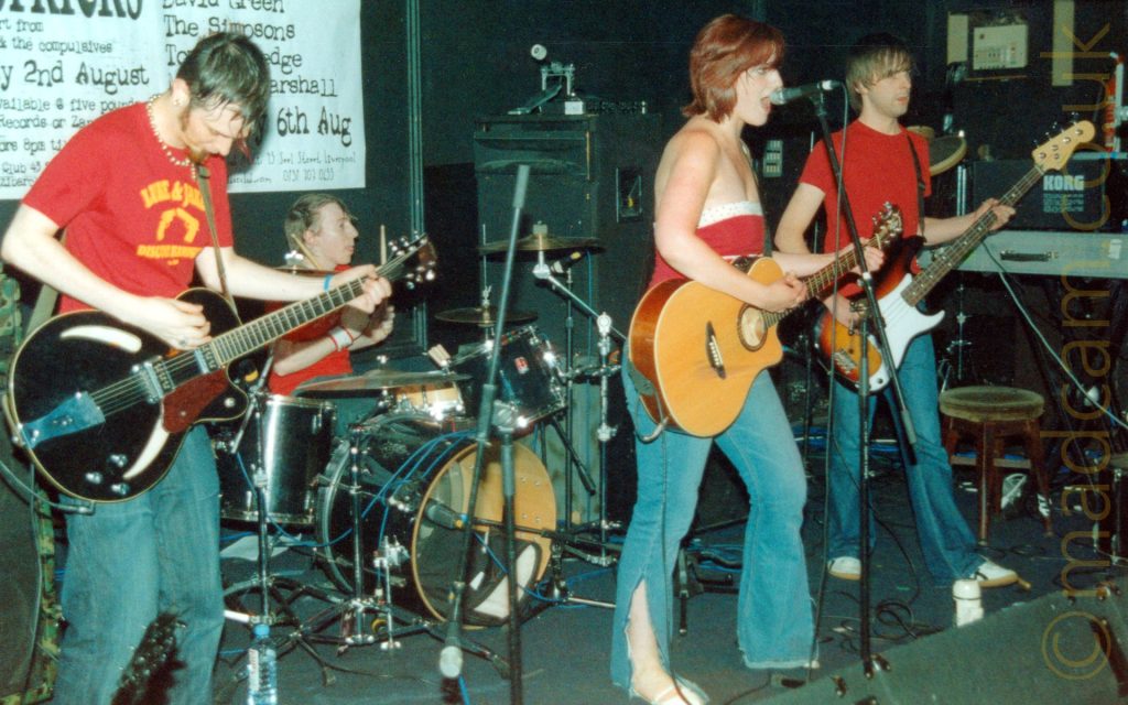 4 members of a band performing on stage. On the left is a man with short dark hair that is longer on top, with ginger sideburns and goatee, wearing a red t-shirt with a yellow motif on the chest, and a blue pair of jeans. He is looking down at a black electric guitar he is holding in his hands. Next is a man with short-ish dark hair, wearing a red t-shirt and blue jeans, sitting behind a black drumkit, holding a pair of drumsticks in his hands, the sticks raised almost vertically. Towards the right of the frame, a woman with shoulder length red hair and wearing a strapless red top with a white band at the top is playing a brown acoustic guitar and singing into a microphone on a stand in front of her. On the left, a man with shaggy brown hair, wearing a plain red t-shirt and a blue pair of jeans is playing a brown and white bass guitar is staring off towards the back of the room. Black wall line the background, with a large white poster with black text advertising events on 2nd and 6th August hanging on the wall on the left.