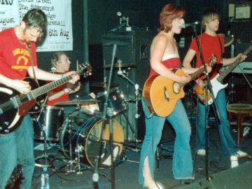 4 members of a band performing on stage. On the left is a man with short dark hair that is longer on top, with ginger sideburns and goatee, wearing a red t-shirt with a yellow motif on the chest, and a blue pair of jeans. He is looking down at a black electric guitar he is holding in his hands. Next is a man with short-ish dark hair, wearing a red t-shirt and blue jeans, sitting behind a black drumkit, holding a pair of drumsticks in his hands, the sticks raised almost vertically. Towards the right of the frame, a woman with shoulder length red hair and wearing a strapless red top with a white band at the top is playing a brown acoustic guitar and singing into a microphone on a stand in front of her. On the left, a man with shaggy brown hair, wearing a plain red t-shirt and a blue pair of jeans is playing a brown and white bass guitar is staring off towards the back of the room. Black wall line the background, with a large white poster with black text advertising events on 2nd and 6th August hanging on the wall on the left.
