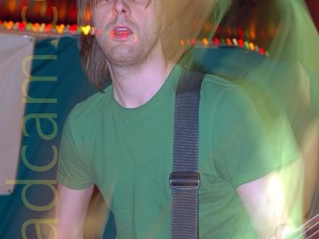 A man with long brown hair, wearing a green t-shirt and a blue pair of jeans, with a black guitar strap slung over his left shoulder attached to the brown bass guitar he is playing. A camera flash has frozen him in place, while a long exposure, a snap zoom, and his movement create secondary images around him. The background has a green and white cloth cover on the wall, with a brown wooden ceiling.