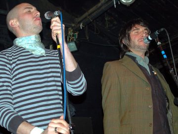 Two men standing on a stage, performing as part of a band. On the left is a man with a shaven head, wearing a grey and black striped, long sleeved t-shirt with a grey cravat round his neck, holding a microphone in his left hand and holding on to the mic stand with his right hand. On the right is a man with shaggy dark hair and a lightly-stubbly beard, wearing a green tweed jacket over a black shirt and a grey neckerchief, singing into a microphone on a stand in front of him. Black walls and ceiling fill the frame behind them.