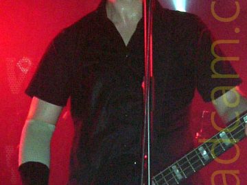A man with close-cropped dark hair, wearing a black shirt and with his elbows and wrists strapped up, playing a black bass guitar and standing, grimacing, behind a microphone on a stand. Red light floods the background, with a single white light shining upwards behind his left shoulder. The head of a fan watching the gig is right at the bottom of the frame.