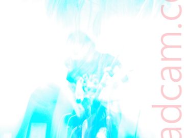 A massively over-exposed long exposure mixed with a snap zoom and an explosion of stage lights, leaving only a vague impression of what is actually there. It starts with a close-up of the head and upper body of a man with shoulder-length hair and wearing a dark t-shirt with a white text logo. His face and most of the background are blown out, massively over-exposed, along with the logo on the t-shirt, while dark areas of the image (his hair, t-shirt, and part of the ceiling behind him) are a light blue. A snap zoom out has a full length view of him, slightly hunched over, playing a guitar. Again, all details have been lost, blown out by the exposure, only blue shadows remaining.