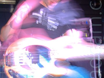 A man on stage, with short dark hair, wearing a black t-shirt with an indistinct white logo and a blue pair of jeans, playing a white bass guitar. A camera flash has frozen him in place, whileca long exposure combined either flashing stage lights and his movement have created purple secondary images around his head and torso. Racks of amplifiers and speakers can be seen in the background, against black walls.