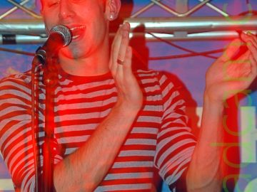 A man with dark, close-cropped hair and wearing a long-sleeved t-shirt with black and white bands around the body, with the sleeves rolled up to his elbows. He is singing into a microphone which is mounted on a stand in front of him, with his hands raised to hes left side as if clapping. The stage lights are casting a red aura around him, while the walls behind him are blue. A tubular metal framework can be seen stretching across the frame in the background.