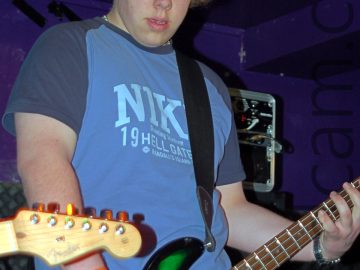 A man with shaggy blond hair, wearing a blue-grey t-shirt with a large Nike logo across the chest and dark grey sleaves, and a blue pair of jeans, and playing an orange and white bass guitar, while looking down at the floor. Various black amplifiers fill the background at the bottom of the frame, with purple walls and ceiling filling the rest.