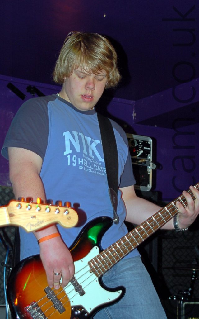 A man with shaggy blond hair, wearing a blue-grey t-shirt with a large Nike logo across the chest and dark grey sleaves, and a blue pair of jeans, and playing an orange and white bass guitar, while looking down at the floor. Various black amplifiers fill the background at the bottom of the frame, with purple walls and ceiling filling the rest.