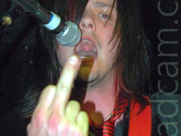 Close-up of the head and upper body of a man with long dark hair covering his right eye, wearing a grey t-shirt, a black waistcoat, and a red tie with thin black stripes, holding up an extended middle finger on his right hand to the camera, with his mouth open, about to stick his tongue out. A red guitar strap is slung over his left shoulder, and there is a microphone on a stand by his mouth on the left of the frame. Black walls and ceiling fill the rest of the frame.