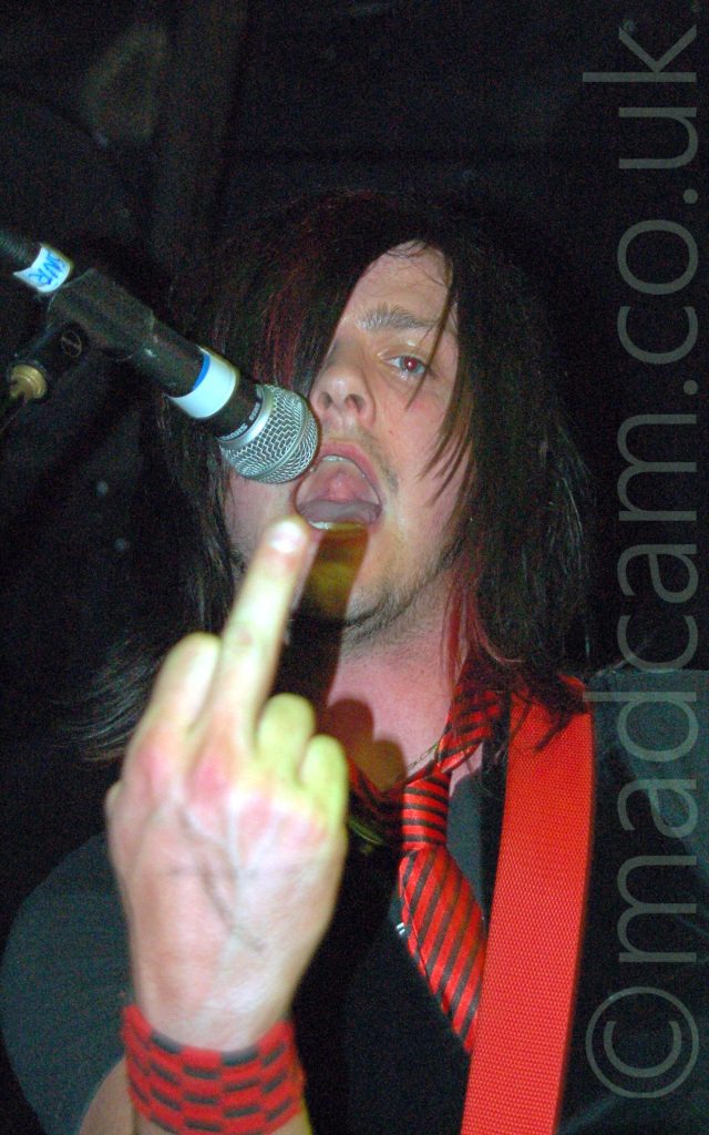 Close-up of the head and upper body of a man with long dark hair covering his right eye, wearing a grey t-shirt, a black waistcoat, and a red tie with thin black stripes, holding up an extended middle finger on his right hand to the camera, with his mouth open, about to stick his tongue out. A red guitar strap is slung over his left shoulder, and there is a microphone on a stand by his mouth on the left of the frame. Black walls and ceiling fill the rest of the frame.