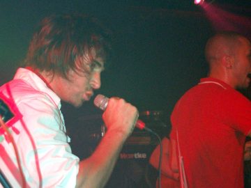 2 men, members of a band, performing on a stage, both fitting to the right. Nearest the camera, on the left of the frame, is a man with long, shaggy hair, wearing a white shirt, and singing into a microphone held in his right hand. On the right is a shaven-headed man wearing a red t-shirt with white piping, singing into a microphone in his right hand. A single red light in the top right is shining towards the front of the stage. Amplifiers line the black walls in the background.