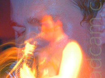 A long exposure snap-zoom shot of a man with short dark hair and wearing a dark tank-top, playing a guitar. Starting as a close-up of his face, a camera flash freezes him in place, while a snap-zoom out while the shutter is open creates a secondary image of him from mid-thigh up, bathed in golden light, with streaks of light floating through the frame.