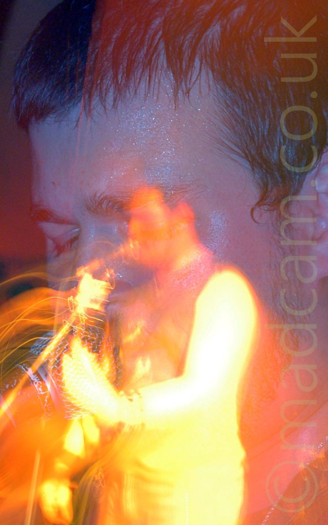 A long exposure snap-zoom shot of a man with short dark hair and wearing a dark tank-top, playing a guitar. Starting as a close-up of his face, a camera flash freezes him in place, while a snap-zoom out while the shutter is open creates a secondary image of him from mid-thigh up, bathed in golden light, with streaks of light floating through the frame.