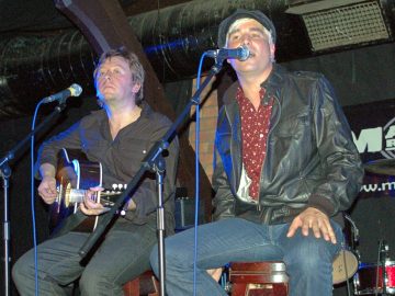 Two men sitting on stools, performing as part of a band on a stage. On the left is a man with shaggy dark hair, wearing a black shirt and black jeans, and playing a dark brown acoustic guitar, with a microphone on a stand in front of him. On the right is a mans with short blond hair, wearing a black corduroy cap, a black leather jacket over a dark red shirt with white and orange flecks, and a blue pair of jeans. He is singing into a microphone on a stand in front of his face. Behind them, black cloths cover brown brick walls, , with thick, dark brown wooden joists holding up the ceiling. A thick, tubular, black ventilation duct runs the width of the frame right at the top.