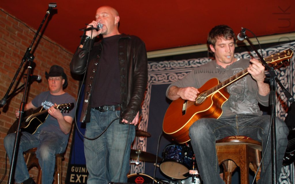 3 members of a band on a stage. On the left is a man wearing a black stetson, a grey t-shirt with a white logo, and a blue pair of jeans, sitting on a stool and playing a black acoustic guitar. In the middle is a man with a shaven head and a thin goatee beard, wearing a leather jacket over a black t-shirt and a blue pair of jeans. He is holding a microphone in his right hand, close to his mouth as he sings in to it, holding the cord in his left hand down by his waist, with his eyes closed. On the right is a man with dark hair, wearing a grey, long-sleeved t-shirt and grey jeans, sitting on a wooden stool and playing a brown acoustic guitar, with a guitar pick held in his lips. A blue cloth with a white knotted design around the edges covers the wall behind them, with a red brick wall on the left, with a red ceiling above them.