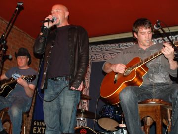 3 members of a band on a stage. On the left is a man wearing a black stetson, a grey t-shirt with a white logo, and a blue pair of jeans, sitting on a stool and playing a black acoustic guitar. In the middle is a man with a shaven head and a thin goatee beard, wearing a leather jacket over a black t-shirt and a blue pair of jeans. He is holding a microphone in his right hand, close to his mouth as he sings in to it, holding the cord in his left hand down by his waist, with his eyes closed. On the right is a man with dark hair, wearing a grey, long-sleeved t-shirt and grey jeans, sitting on a wooden stool and playing a brown acoustic guitar, with a guitar pick held in his lips. A blue cloth with a white knotted design around the edges covers the wall behind them, with a red brick wall on the left, with a red ceiling above them.