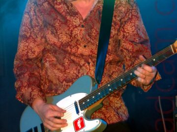 A man on stage wearing a black stetson, a red shirt with an orange flowery print, and a dark-coloured pair of trousers, playing a grey and white electric guitar with the letters "ZU" marked out in tape and a red square logo of a white stick figure holding a guitar with it's left hand raised in the air. A beam of blue light shines down behind him, illuminating the rear of the stage, diffused through a cloud of smoke.