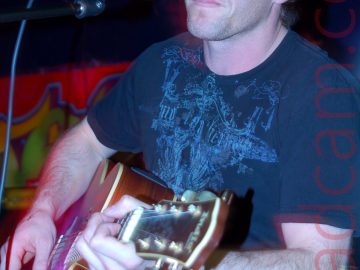 A man with dark hair and wearing a dark blue t-shirt and blue jeans, sitting on a stool and playing a brown acoustic guitar, while staring off into the distance. A microphone on a stand is positioned in front of him, but he isn't singing. A red aura from the stage lights is visible around his arms and head. The walls behind him are black, with a red poster with indecipherable yellow and green writing on the left of the frame.
