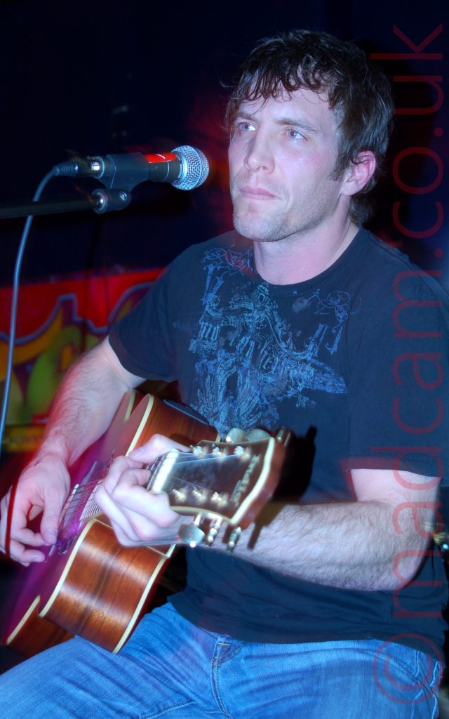 A man with dark hair and wearing a dark blue t-shirt and blue jeans, sitting on a stool and playing a brown acoustic guitar, while staring off into the distance. A microphone on a stand is positioned in front of him, but he isn't singing. A red aura from the stage lights is visible around his arms and head. The walls behind him are black, with a red poster with indecipherable yellow and green writing on the left of the frame.