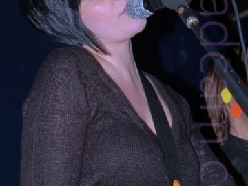 A woman with chin-length black hair cut in a pixie bob, singing into a microphone mounted on a stand in front of her. Her head is tilted slightly to her right, and her eyes are slightly closed. She is wearing a long-sleeved black woollen dress, with a wide red leather belt tied around her waist. A black guitar strap is slung over her left shoulder, attached to a white guitar sneaking into shot at the bottom of the frame. The head and shoulder of a person with shoulder-length blond hair is visible next to her, on the right of the frame, behind the mic stand. Dark blue walls and black ceiling fill the rest of the frame.
