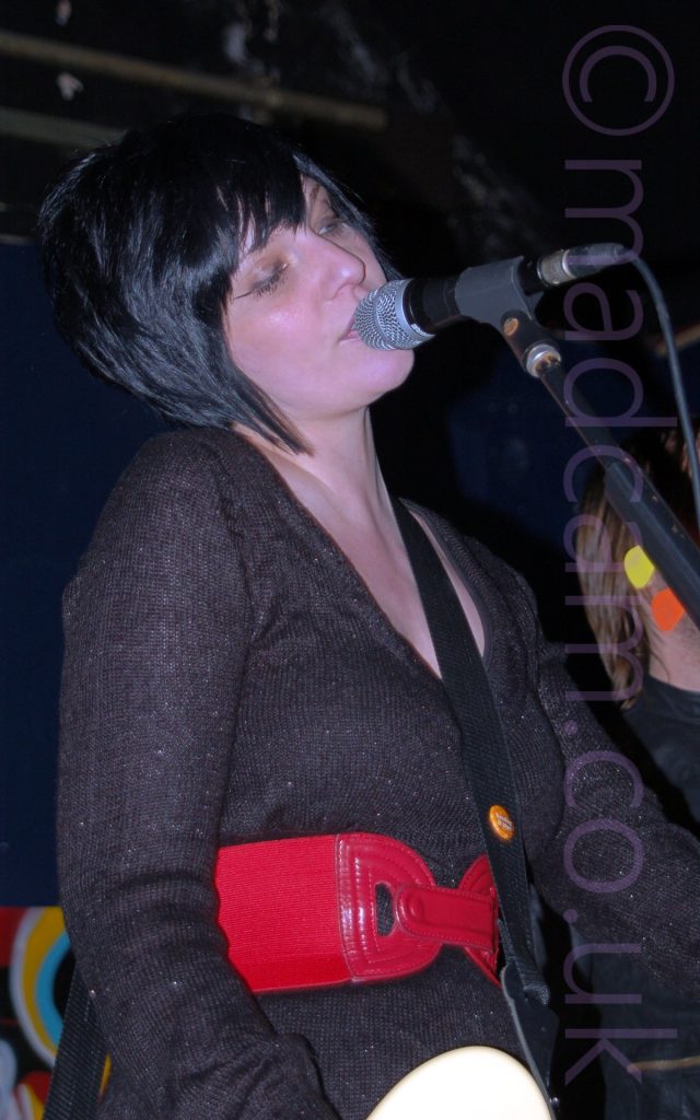 A woman with chin-length black hair cut in a pixie bob, singing into a microphone mounted on a stand in front of her. Her head is tilted slightly to her right, and her eyes are slightly closed. She is wearing a long-sleeved black woollen dress, with a wide red leather belt tied around her waist. A black guitar strap is slung over her left shoulder, attached to a white guitar sneaking into shot at the bottom of the frame. The head and shoulder of a person with shoulder-length blond hair is visible next to her, on the right of the frame, behind the mic stand. Dark blue walls and black ceiling fill the rest of the frame.