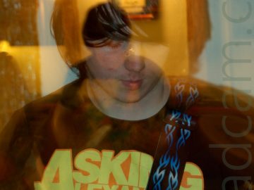 A young man with long-ish balck hair, wearing a black t-shirt with a yellow, blue, and orange logo of a many-eyed, blue, demonic figure and yellow text "Asking Alexandria", and ith a black guitar strap with blue flame markings slung over his left shoulder. He is looking down at the ground in front of him. A camera flash has frozen him in place, while a long exposure, a snap-zoom, and his movement have created secondary images, lit with yellow light.White walls fill the background, with a framed picture appearing in 2 different places, and wall-mounted lights that leave smears of light across the frame.