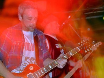 A man with short, close-cropped greying hair and beard, and wearing an open blue, red, and white checked shirt over a white t-shirt, playing an orange electric guitar. A camera flash has frozen him in place, while a long exposure and a slow zoom in has combined with the stage lights to paint the area around him with red and yellow light. In the background, a man wearing a blue cap and wearing a red and white basketball shirt and playing a bass guitar is singing into a microphone. Black walls mark the edges of the room, with a bright light smeared into a streak in the top right corner.