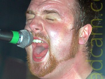 Close-up, head and shoulders shot of a man with short dark hair and a ginger goatee beard and sideburns, wearing a black t-shirt. He is singing in to a microphone on a stand in front of him. Stage lights behind him are casting a slight green halo around the back of his head.