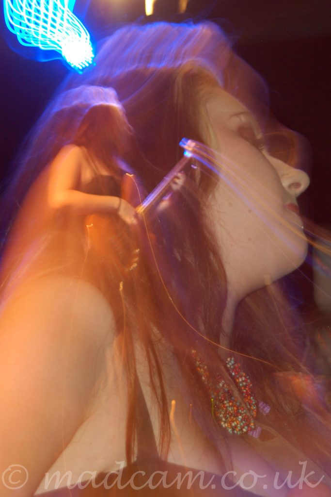 A woman with long dark hair tumbling over her shoulders, wearing a strappy black dress that leaves her shoulders and arms bare, and playing a red electric bass guitar. Her head is turned to the left, and tilted slightly back. A long exposure combined with a snap zoom results in 2 separate but connected images, a close-up of her head and shoulders on the right, and a distant, full-length shot on the left, both with a golden cast. Streaks of purple and blue light surround her head, with black walls and ceiling in the background.