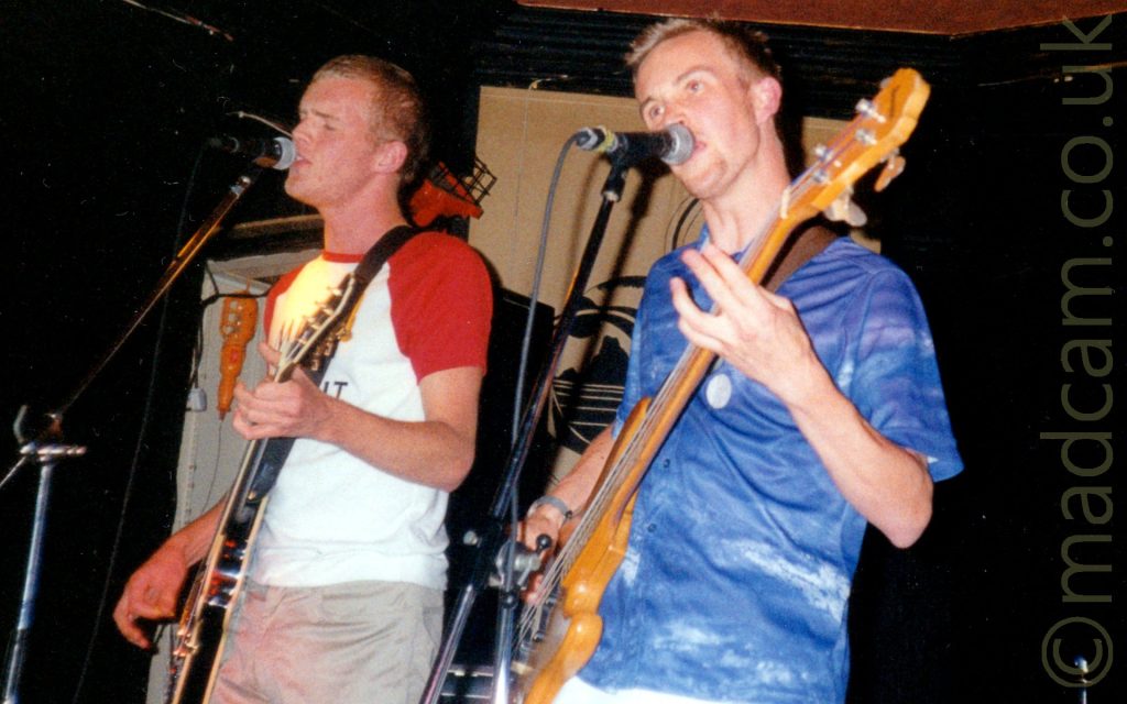2 men, members of a rock band, performing on a stage. On the left is a man with short blond hair, wearing a white t-shirt with red sleeves, and grey trousers, playing a brown electric guitar and singing into a microphone on a stand in front of his face. On the right is a man with short blond hair, wearing a blue shirt, and playing a brown bass guitar, while singing into a mic on a stand in front of him. Black walls and ceiling fill the rest of the room.