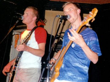 2 men, members of a rock band, performing on a stage. On the left is a man with short blond hair, wearing a white t-shirt with red sleeves, and grey trousers, playing a brown electric guitar and singing into a microphone on a stand in front of his face. On the right is a man with short blond hair, wearing a blue shirt, and playing a brown bass guitar, while singing into a mic on a stand in front of him. Black walls and ceiling fill the rest of the room.