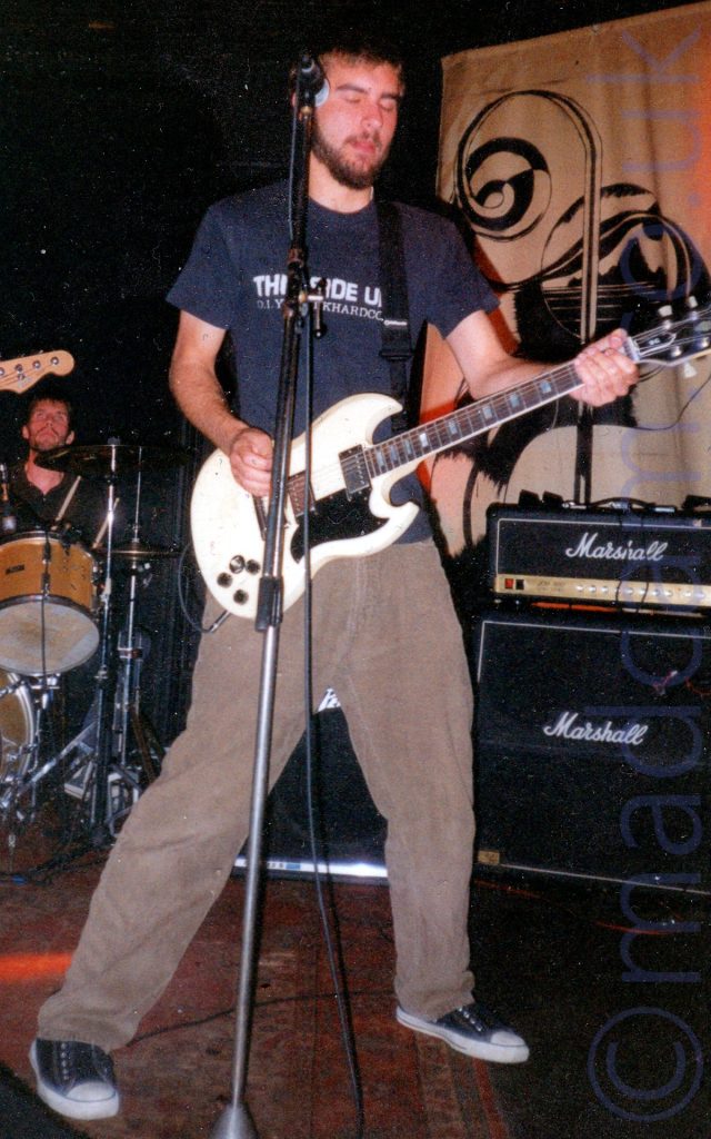 A man with short dark hair and a short beard, wearing a dark blue t-shirt with an indistinct white logo, and brown trousers, playing a white electric guitar with his eyes closed, facing to the right. A microphone on a stand is positioned slightly off to his left. A man is sitting behind a brown drum kit in the background on the left, and appears to be looking up at the head of a guitar that appears to be floating in mid-air. A black Marshall amplifier is on the right of the frame, under a black mural on a brown piece of cloth mounted on the black wall.