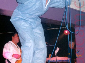 A man with very short dark hair, wearing a blue coverall and blue trainers, standing on a red metal railing at the front of a stage and screaming in to a microphone held in his left hand, with a blue lead attached to the mic and looped around his hand. He is crouched over slightly, as the ceiling is a bit too low for him to stand erect. Purple walls and white ceiling stretch aross the background behind him. In a recess in the purple wall, a man in similar white coveralls has a brown guitar slung over his shoulder with a black strap.
