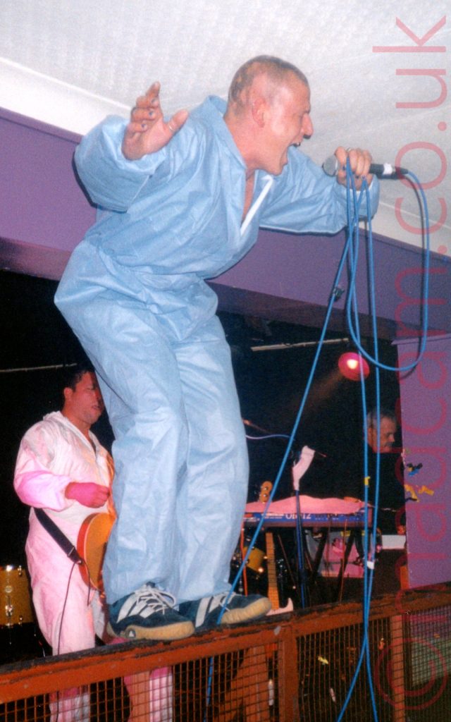A man with very short dark hair, wearing a blue coverall and blue trainers, standing on a red metal railing at the front of a stage and screaming in to a microphone held in his left hand, with a blue lead attached to the mic and looped around his hand. He is crouched over slightly, as the ceiling is a bit too low for him to stand erect. Purple walls and white ceiling stretch aross the background behind him. In a recess in the purple wall, a man in similar white coveralls has a brown guitar slung over his shoulder with a black strap.