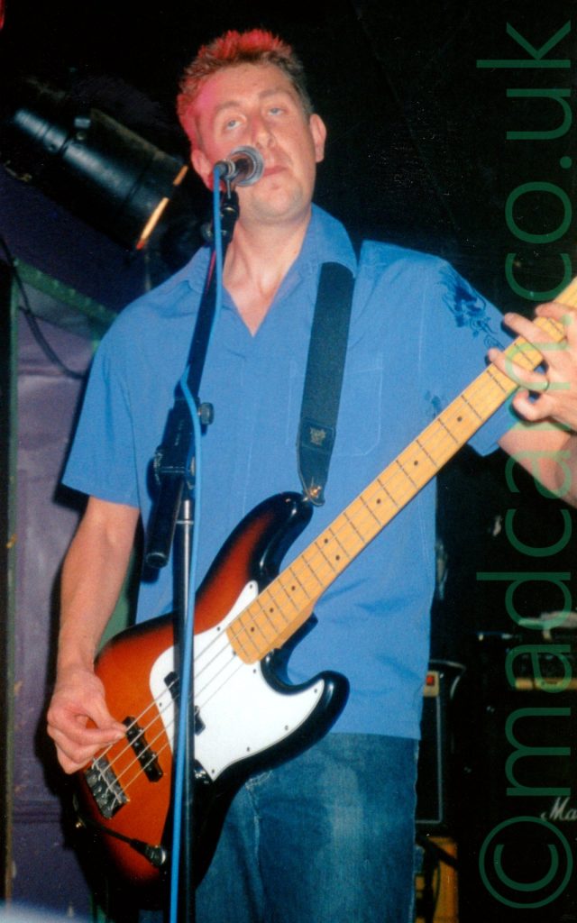 A man with short brown hair, wearing a blue shirt and a blue pair of jeaans, with a brown and white slung over his left shoulder by a black strap, pulling faces into a microphone on a stand in front of him. Black walls and ceiling fill the rest of he frame behind him.