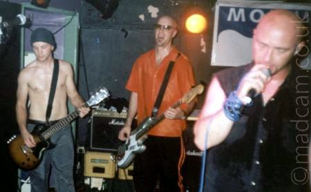 3 members of a band performing on stage - at the back on the left is a man wearing a beanie hat, and a pair of grey trousers, and not much else, playing a brown electric guitar, and sweating profusely. In the middle is a man with a shaven head and wearing a pair of pair of sunglasses, with a red shir and black trousers, playing a grey and white bass guitar. On the right, in the foreground, is a man with a shaven head, wearing a sleeveless dark shirt, holding a microphone to his mouth, with the cord for the mic wrapped several times around his wrist. Black walls and ceiling fill the background, with an orange light in the middle shining directly into the camera.