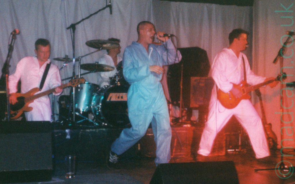 4 members of a rock band on stage, all dressed in white coveralls, except the singer, who is in blue. On the left is a man with short dark hair, playing a brown bass guitar. In the middle, at the back, is a man sitting behind a metallic blue drumkit. At the front is a man with very short dark hair, holding a microphone in his left hand as he sings in to it. On the right is a man with short dark hair, smoking a cigarette as he plays a brown electric guitar. Grey curtains cover the walls in the background.
