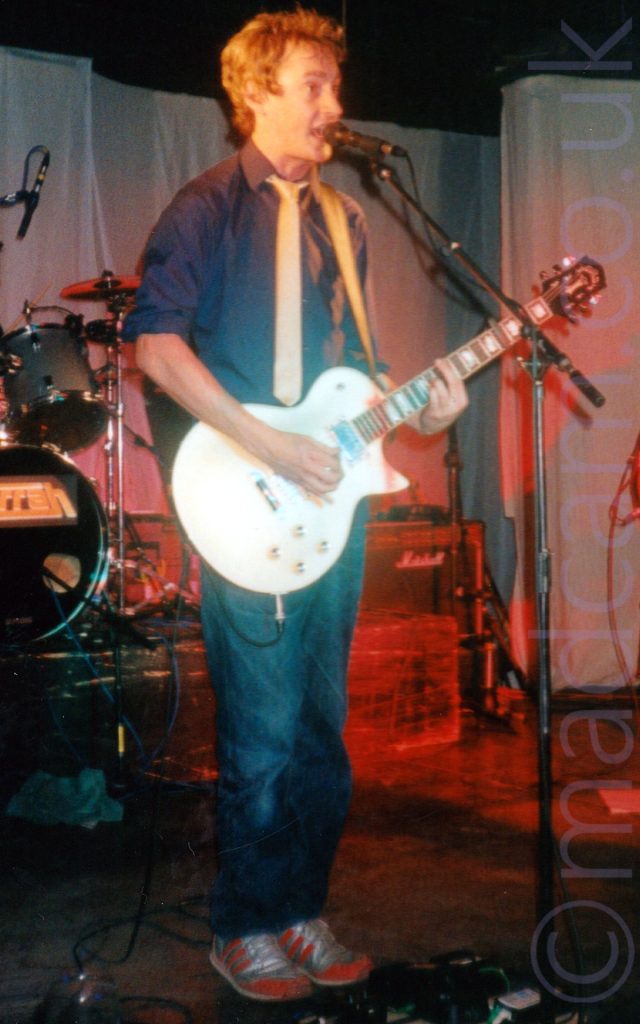 A man performing on stage. He has short, slightly curly brown hair, wearing a black shirt with a gold tie, blue jeans, and red and grey trainers, playing a white electric guitar while singing into a microphone on a stand in front of him. Part of a drum kit can be seen in the background on the left, with grey curtains covering the walls, and black ceiling. Parts of the curtain and some amps at the back of the stage are coloured red because of some stage lights.