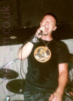 A man with a shaved head on a stage. He is wearing a black t-shirt with a yellow and green circular logo and dark trousers, and has a microphone in his curtains covering the walls at the back of the stage, and a black ceiling.right hand which he is singing in to with his eyes closed, it's lead wrapped around his right wrist. Part of a black drum kit can be seen in the background on the left, with grey curtains covering the walls at the back of the stage, with a black ceiling.