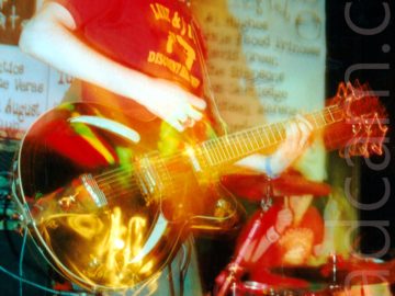 A man with lank dark hair and almost impressive sideburns, along with the beginnings of a goatee beard, wearing a red t-shirt with a yellow text-based logo that is almost indecipherable, and baggy blue jeans. He is playing a brown electric guitar, looking down at his left hand on the strings on the guitars neck while his right hand plays a chord. A long exposure combined with his movement has created some ghostly secondary images, mostly around the body and neck of the guitar, as well as some yellow and white light trails. A person wearing a red t-shirt can be seen sitting behind a drum kit in the background at the bottom right of the frame. A pair of large white posters, with text apparently advertising up-coming events at the venue dominate black walls behind this man.