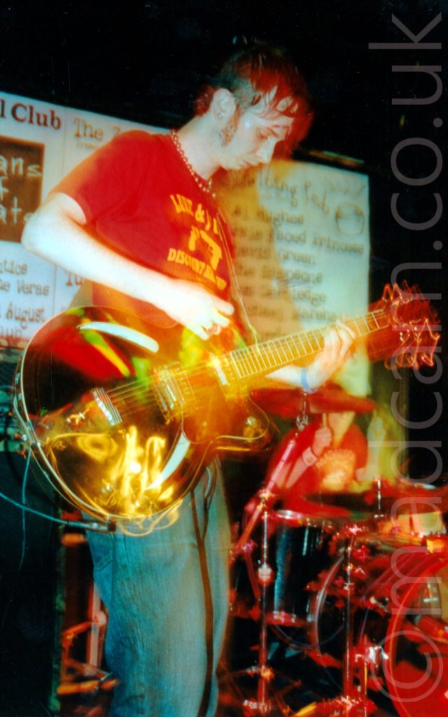 A man with lank dark hair and almost impressive sideburns, along with the beginnings of a goatee beard, wearing a red t-shirt with a yellow text-based logo that is almost indecipherable, and baggy blue jeans. He is playing a brown electric guitar, looking down at his left hand on the strings on the guitars neck while his right hand plays a chord. A long exposure combined with his movement has created some ghostly secondary images, mostly around the body and neck of the guitar, as well as some yellow and white light trails. A person wearing a red t-shirt can be seen sitting behind a drum kit in the background at the bottom right of the frame. A pair of large white posters, with text apparently advertising up-coming events at the venue dominate black walls behind this man.