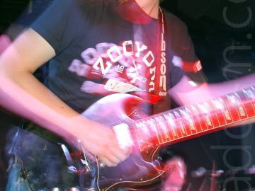 Close-up of a man with shoulder-length wavy brown hair, wearing a blue t-shirt with a white logo and a blue pair of jeans, playing a black electric guitar whikle facing to the right. A long exposure combined with slight camera movements have created purple secondary images, his arms and body appearing almost ghost-like. In the background, a man can be seen playing on a drum kit in the bottom right of the image, with black walls and ceiling filling the rest of the frame.