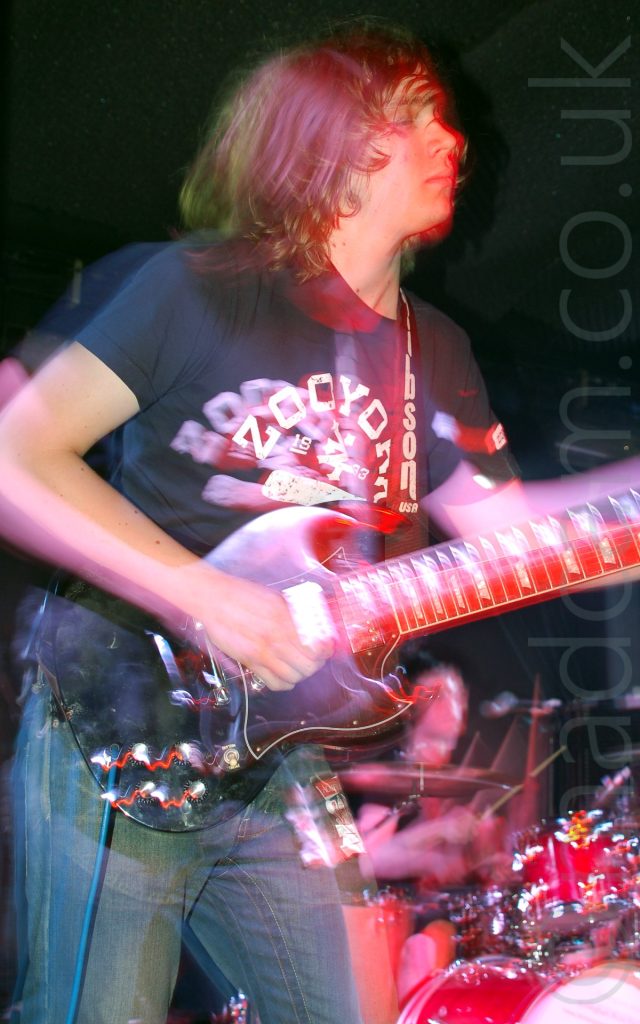 Close-up of a man with shoulder-length wavy brown hair, wearing a blue t-shirt with a white logo and a blue pair of jeans, playing a black electric guitar whikle facing to the right. A long exposure combined with slight camera movements have created purple secondary images, his arms and body appearing almost ghost-like. In the background, a man can be seen playing on a drum kit in the bottom right of the image, with black walls and ceiling filling the rest of the frame.