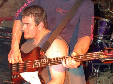 2 members of a band performing on stage, and indulging in some hi-jinks. On the left is a man with short, dark hair and a straggly beard, wearing a grey vest and a blue pair of jeans, crouching down on the floor, holding an electric guitar and facing off to the left. Behind him, on the right, is another man with short dark hair and beard, wearing a blue sweater with red logos on the chest and sides of the arms, and a faded blue pair of jeans. He is holding a red wood and white bass guitar, which is wrapped around the other man. A black and chrome drum kit is in the background on the right, with a brown brick wall filling the rest of the frame.