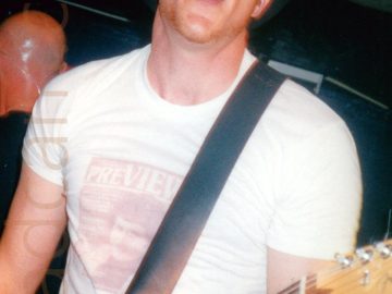 Close-up of a man wearing a black, wide-brimmed hat, and a white t-shirt with a brown image of someone's face below the word "Preview", looking directly at the camera with his mouth open while playing a silvery-grey guitar. A man with a bald head is standing over his shoulder, facing the other way. Black walls and dark ceiling fill the rest of the frame.