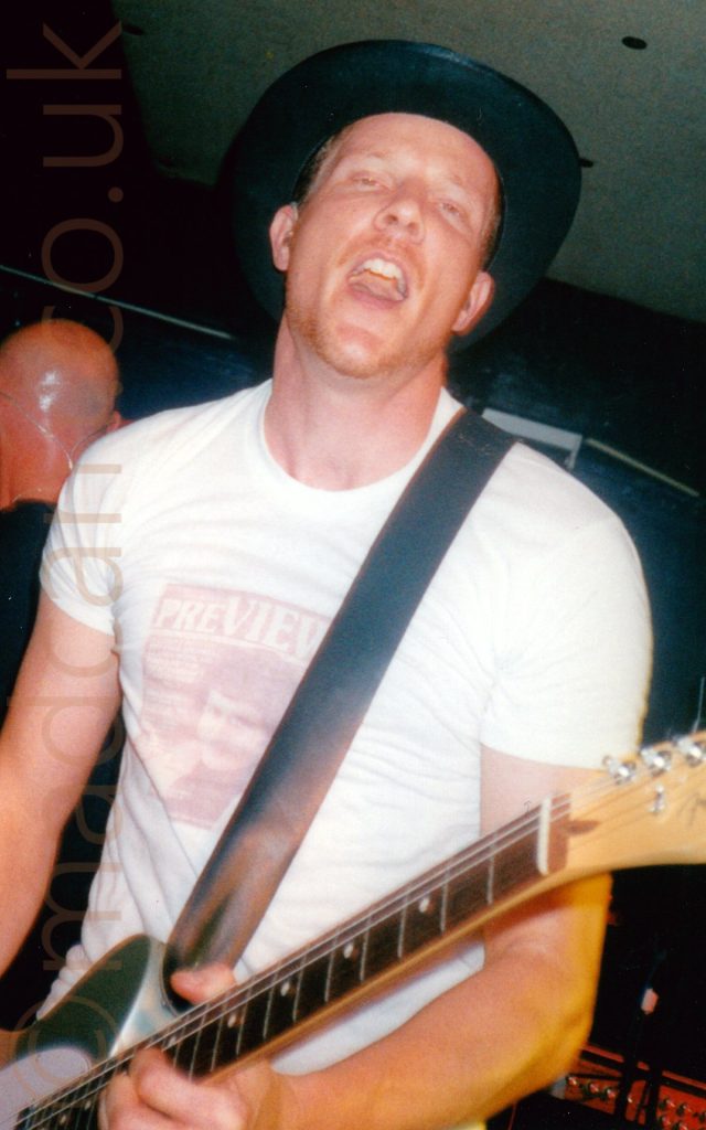 Close-up of a man wearing a black, wide-brimmed hat, and a white t-shirt with a brown image of someone's face below the word "Preview", looking directly at the camera with his mouth open while playing a silvery-grey guitar. A man with a bald head is standing over his shoulder, facing the other way. Black walls and dark ceiling fill the rest of the frame.