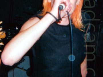 A woman with shoulder length orange hair, wearing a black t-shirt and grey trousers, holding a microphone to her mouth, with the cord for the mic wrapped around her right wrist. She is staring past the camera. A drum kit is visible in the background, with black walls and ceiling filling the rest of the frame.